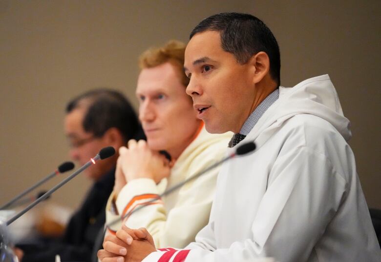 One political leader speaks at a table while two others listen in the background.