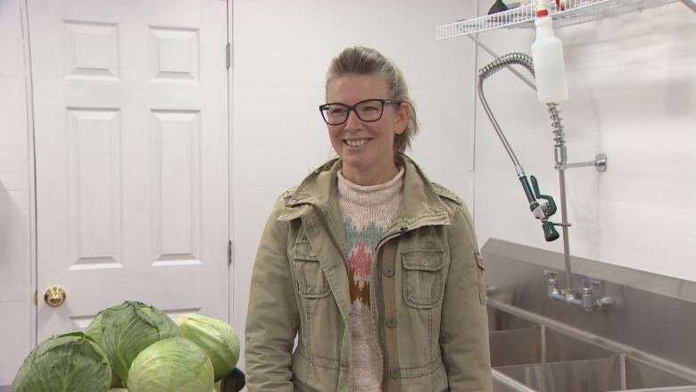 A woman stands in a kitchen next to a bowl of green cabbages.