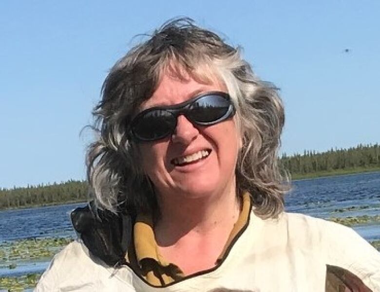 A woman wearing sunglasses smiles for a photo in front of an expanse of water.