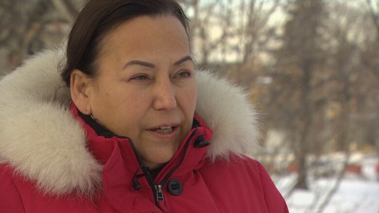 A woman wearing a red parka with a furry trim around the hood speaks outside. Behind here, there are snow and trees.