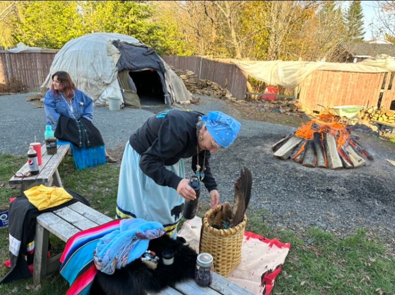 Elder Imelda Perley stands outside, bending over a basket, also called a sacred bundle. Behind her are the sweat lodge and sacred fire.