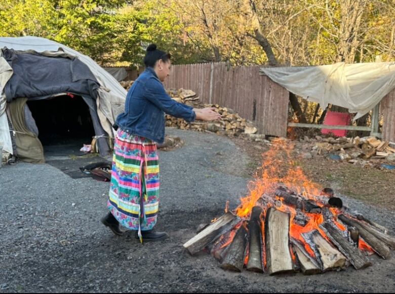 A woman in a denim jacket and long, multi-coloured skirt stands outside, warming her hair over a sacred fire. Behind her is a sweat lodge.