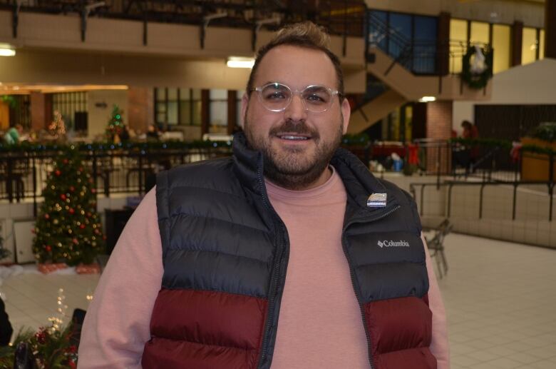 A man poses for a portrait, he's wearing a pink sweatshirt and a black puffer vest. 