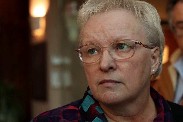 A woman with short grey hair and glasses glances to the left of the photo with a serious expression. It's fairly close-up and only the collar of a black blouse with aqua squares and some red patterning is visible.