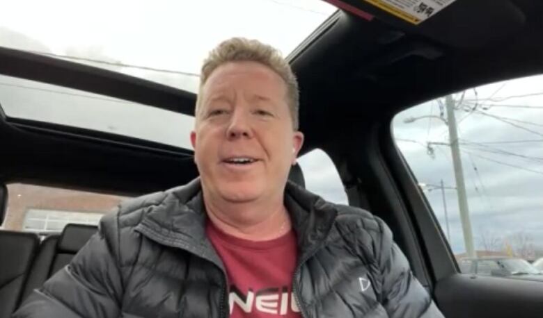 A man in a black coat and red shirt speaks to his phone in his car. 