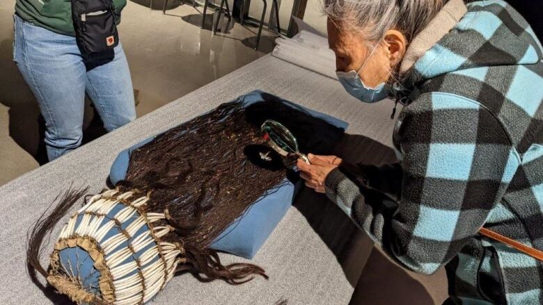 A woman wearing a mask examines an Indigenous headdress with a magnifying glass.