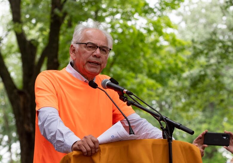 A man stands and speaks at a podium wearing an orange shirt. 