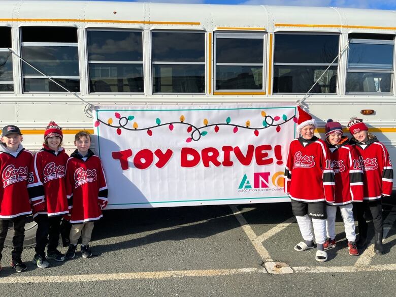 Six young boys stand in front of a white bus with a banner reading 'Toy Drive!' attached to it. They're all wearing red hockey jerseys with black and white trim.