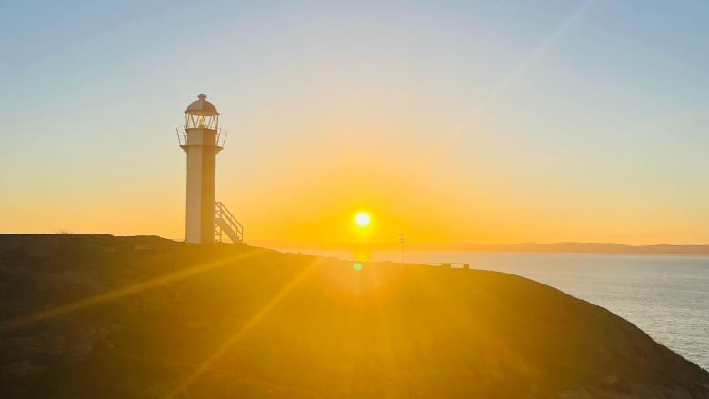 The sun is coming up over the ocean, with a lighthouse on a cliff overhead.