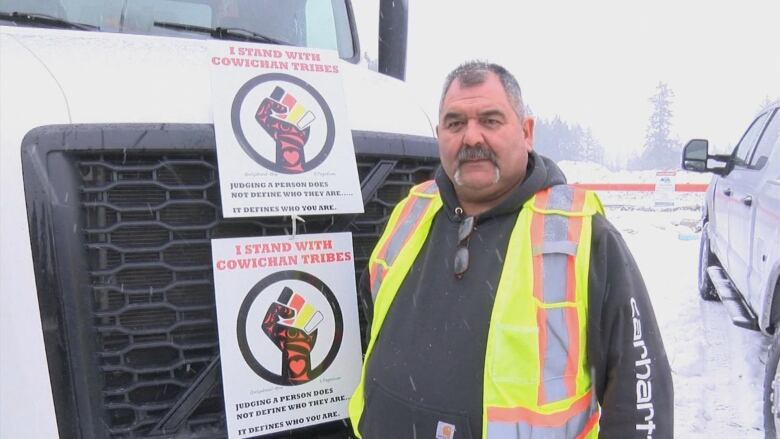 A man in a high-vis vest stands next to a truck and a board that reads 'I stand with Cowichan Tribes'.