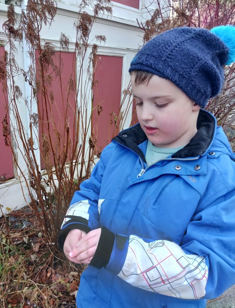 A young boy, dressed in a winter jacket and a hat, is pictured looking down at his hands, in which he is holding something that's hidden from view.