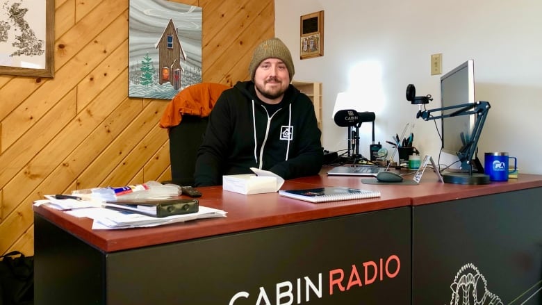 Man sits behind desk in front of computer and microphone, smiles at camera.