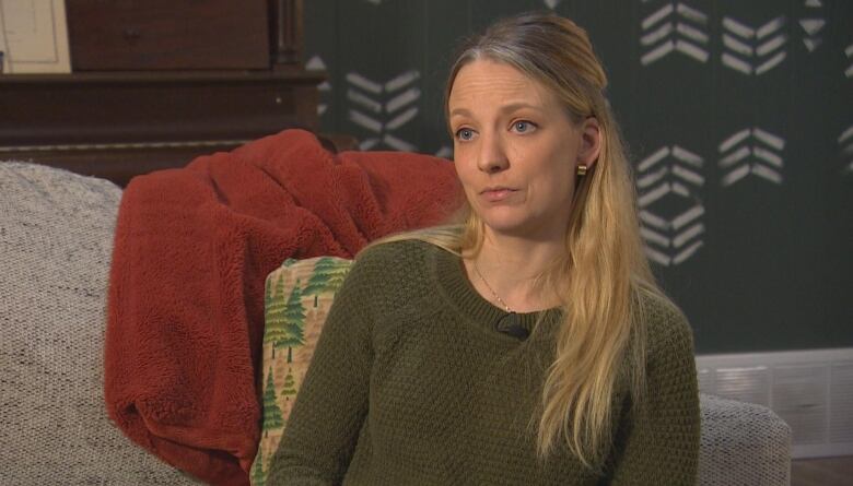 A woman with long blond hair, wearing a green sweater, sits on a couch and looks at a person off-camera with a serious expression.