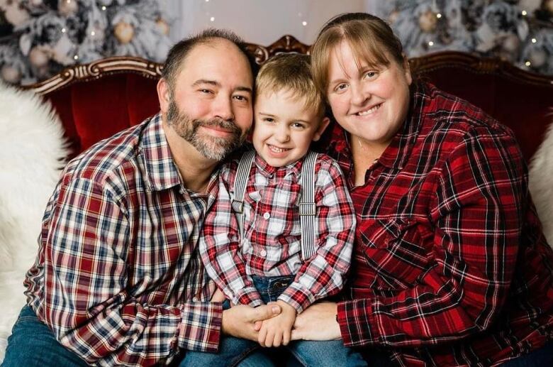 Three people, all wearing red plaid, hug and smile. The person in the middle is a young child.