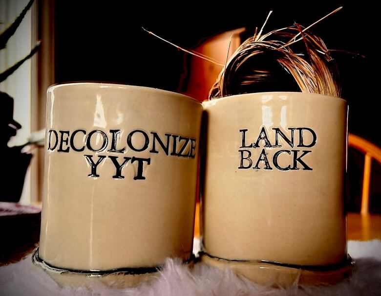 Two glazed clay cups with the words Decolonize YYT and Land Back sit on a table.