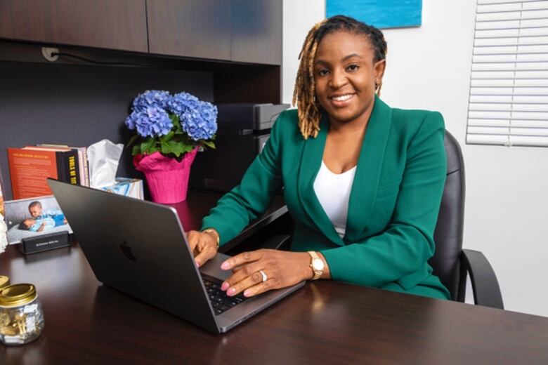 A smiling woman in a green business suit sits at her desk with a laptop.