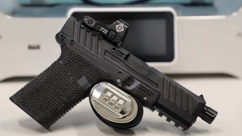 A black handgun sits on a table, in front of a grey printer.