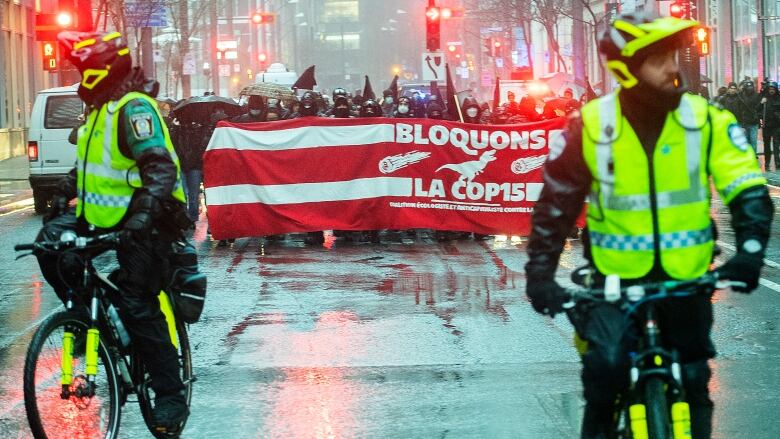 Police on bikes ahead of protesters holding signs in rainy downtown Montreal.