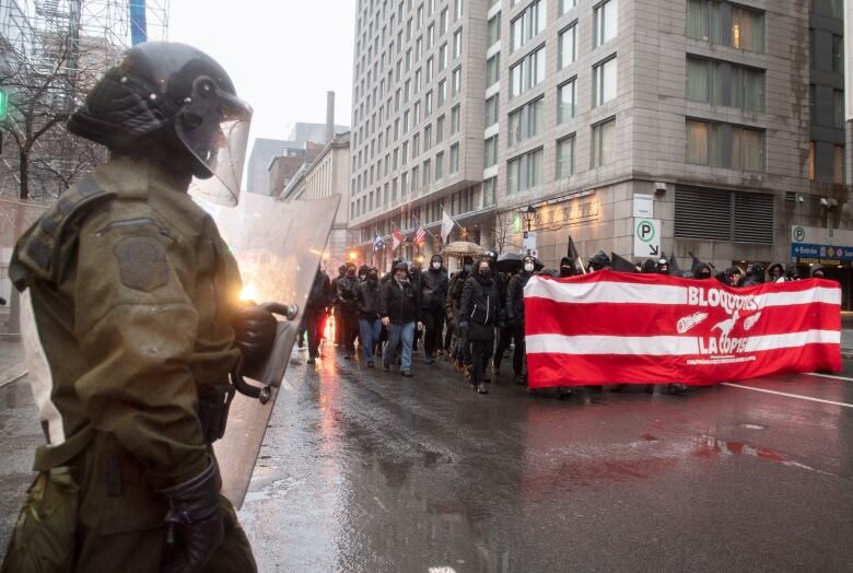 An armed police officer looks on as protesters march with a large red banner.