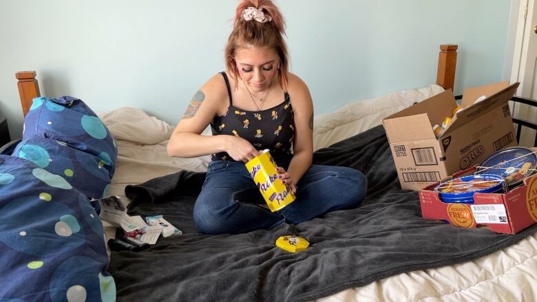 A young woman wearing a black tank top sits on a bed. She is putting something into a yellow tin. There are two cardboard boxes beside her.