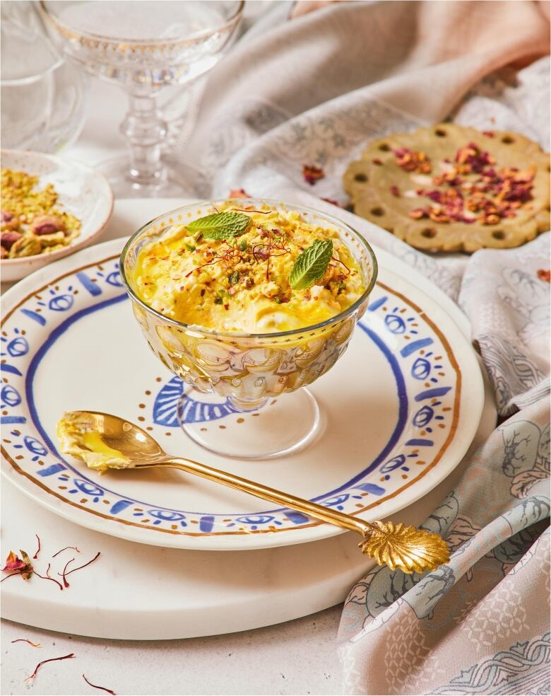 A footed glass bowl with a yogurt dessert in it. It's on a blue and white plate with a gold spoon next to it, and sitting on top of a table with a patterned tablecloth. 