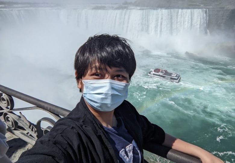 A person stands in front of a waterfall with a boat floating in the background.