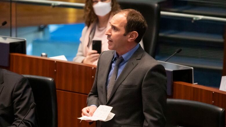 A man in a dark suit speaks on the floor of city council.