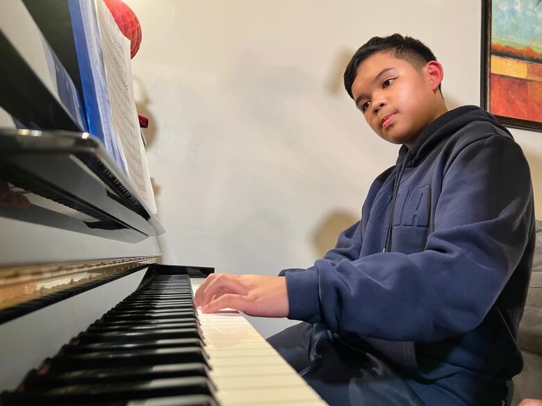 A boy wearing a blue hoodie sits at a piano. His hand is on the keys and he is looking at sheet music in front of him.