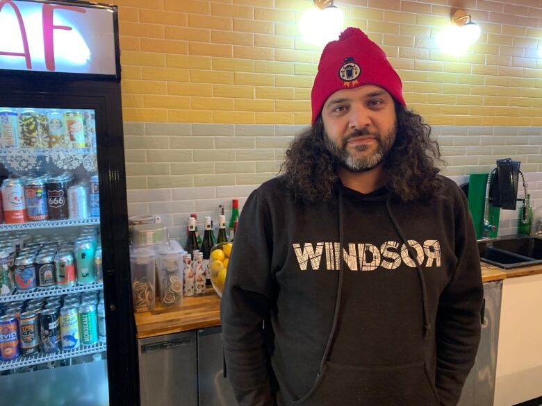 A man stands beside a beer fridge wearing a sweater that says 'Windsor'