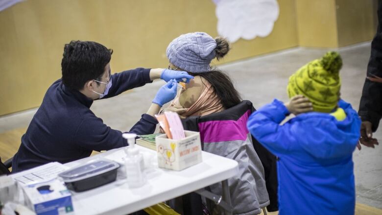 A health worker at a long table covered in vaccination equipment sticks a needle in a child's arm while another child behind them covers their ears. Both children are wearing overcoats.