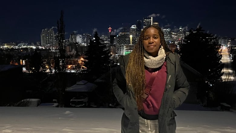 A woman stands in a winter jacket and boots stands in the snow in front of the Calgary skyline at night.