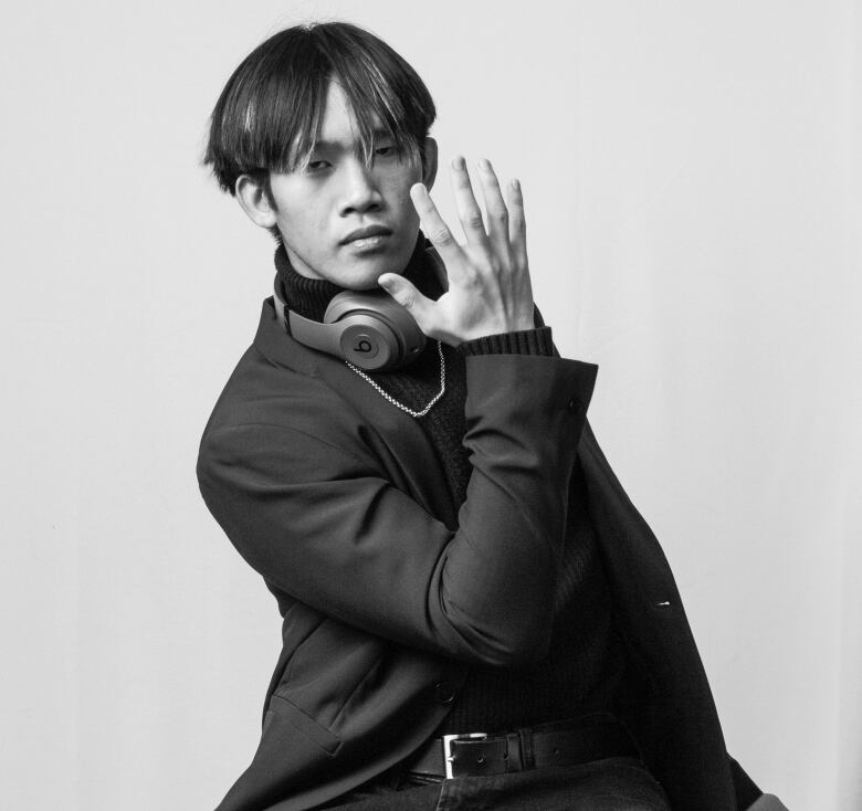 A young man with headphones around his neck sits on a stool, leaning a little to his right, with his right hand held open towards himself, slightly in front of the left side of his face.