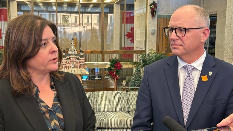 A woman with shoulder length brown hair, wearing a black blazer and dark print blouse, stands next to a man with balding grey hair and glasses, wearing a dark suit. A microphone can be seen in the foreground of the image.