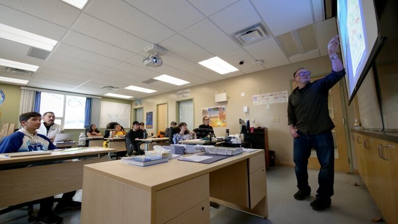 A classroom of students sit at their desks, watching a man with glasses point to a projector screen.