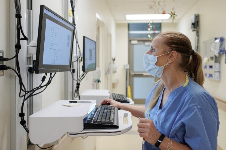 A woman wearing blue hospital scrubs and a medical masks stands in front of a computer monitor in a hospital.