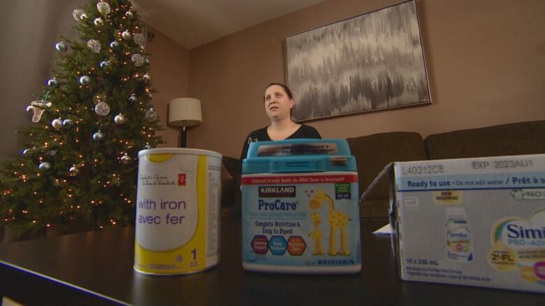 A woman sits on a couch to the right of a lit Christmas tree. Three different kinds of infant formula products are on a coffee table in the foreground. 