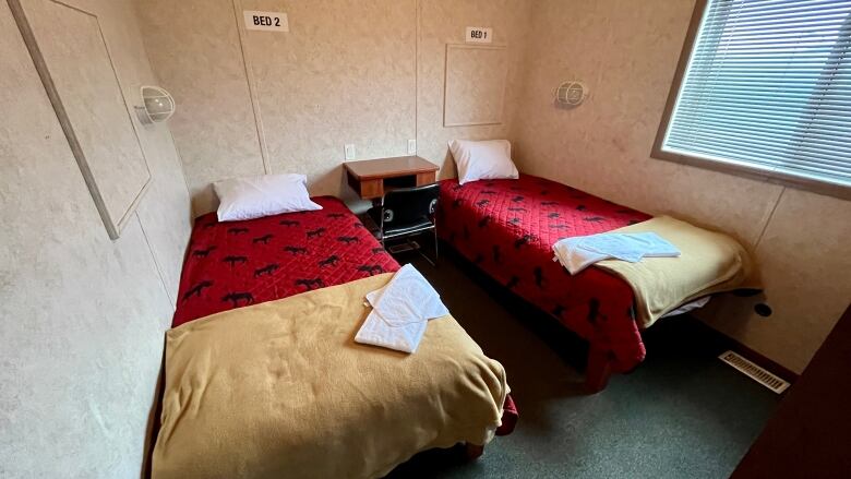 Two single beds side by side with red coverlets with a moose pattern, fleece blankets folded at the bottom, and towels folded on top. There is a window on one wall and a small table between the beds. 