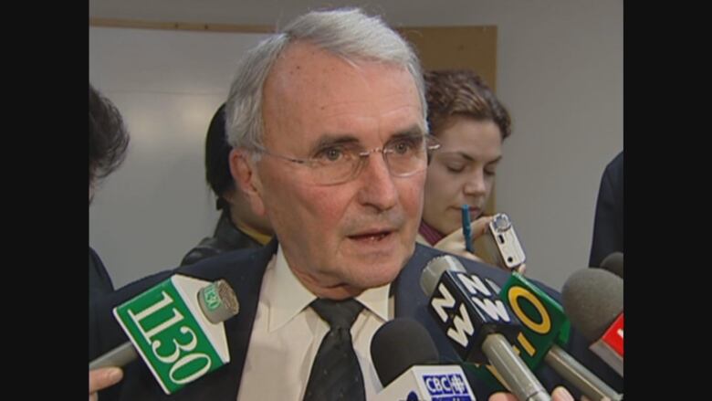 A man with grey hair and glasses speaks into several reporters' microphones