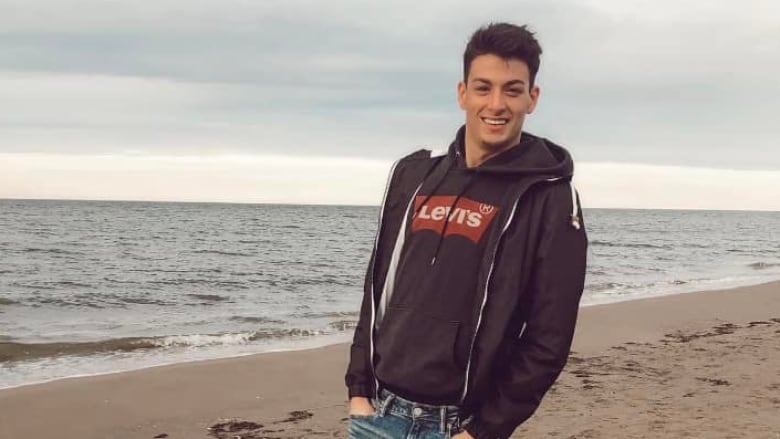 A man with short dark hair stands on a beach with small waves wearing a brown hoodie with his hands in his jean pockets.