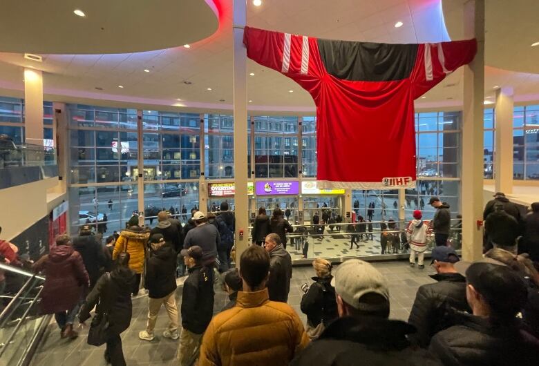 Inside a building with large windows. Lots of people are gathered. A large red jersey hangs from the ceiling.