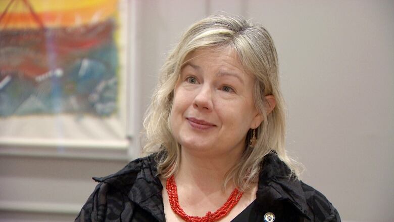 A white woman with a chunky red necklace and shoulder-length blonde hair stands in an office with a colourful art piece visible over her shoulder.