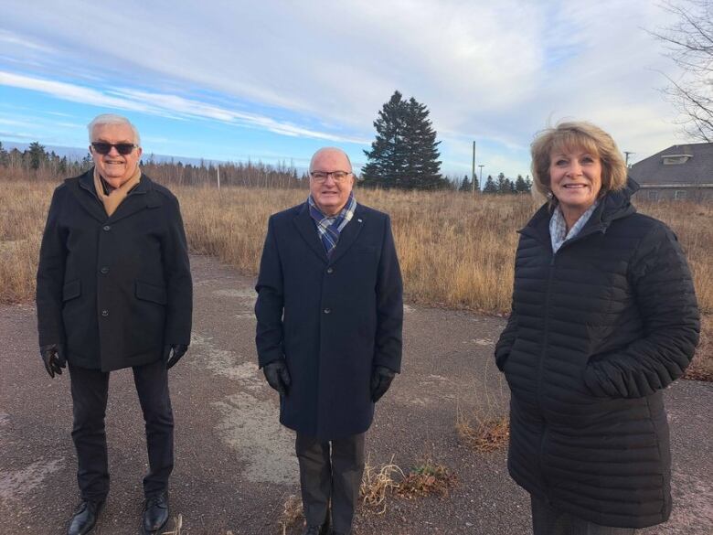 Two men and a woman standing in a gravel lot in front of a field.