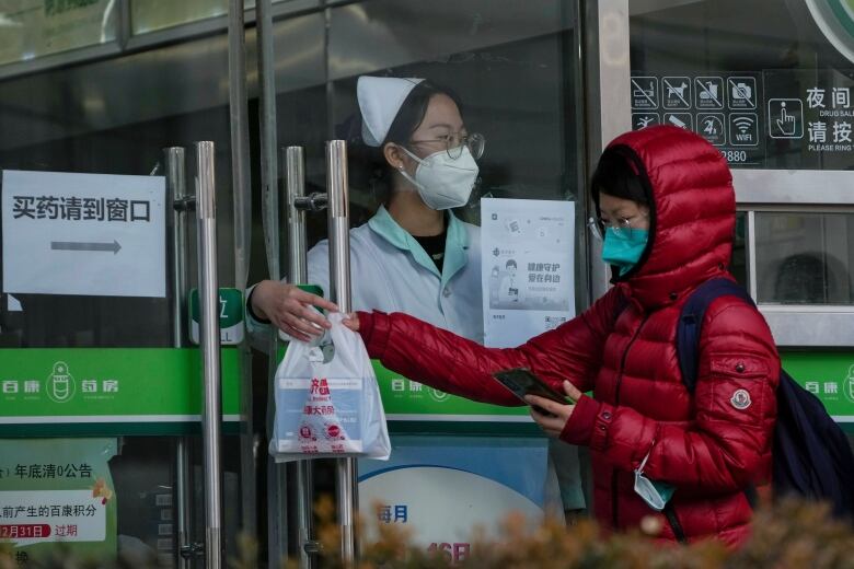 A woman collects COVID-19 antigen kits from a worker through a set of glass doors at a pharmacy in Beijing.