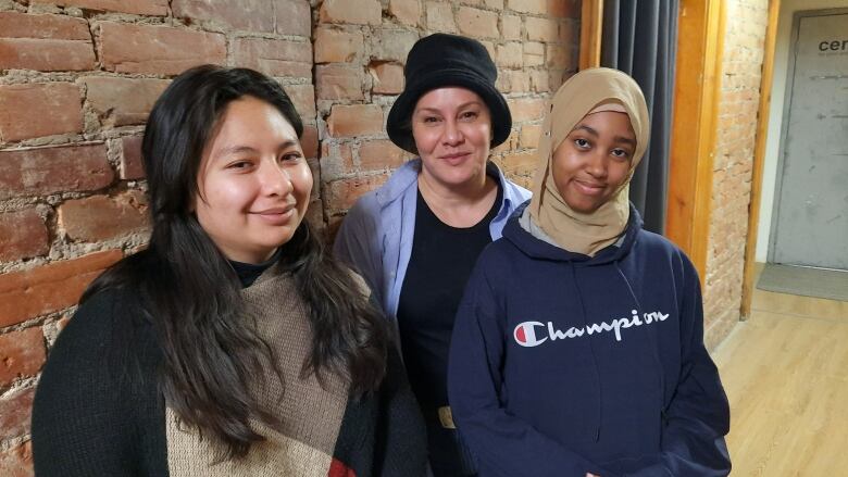 Three women stand in front of a brick wall in a hallway.