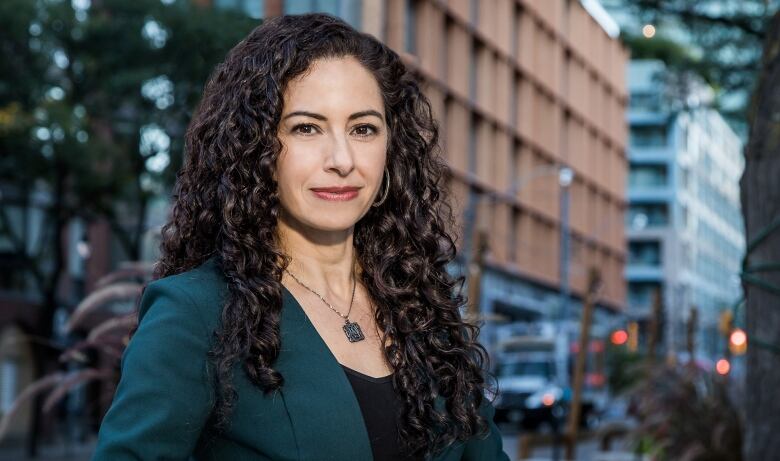 A woman with long, dark curly hair and a teal blazer stands on a street.