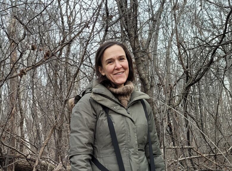 A woman stands in the woods, smiling to the camera.