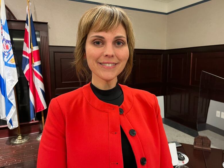 A woman wearing a red coat standing in front of flags inside council chambers. 