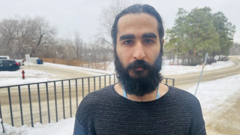 A man with a beard, wearing a black sweater, stands outside in a snowy area, looking at the camera.