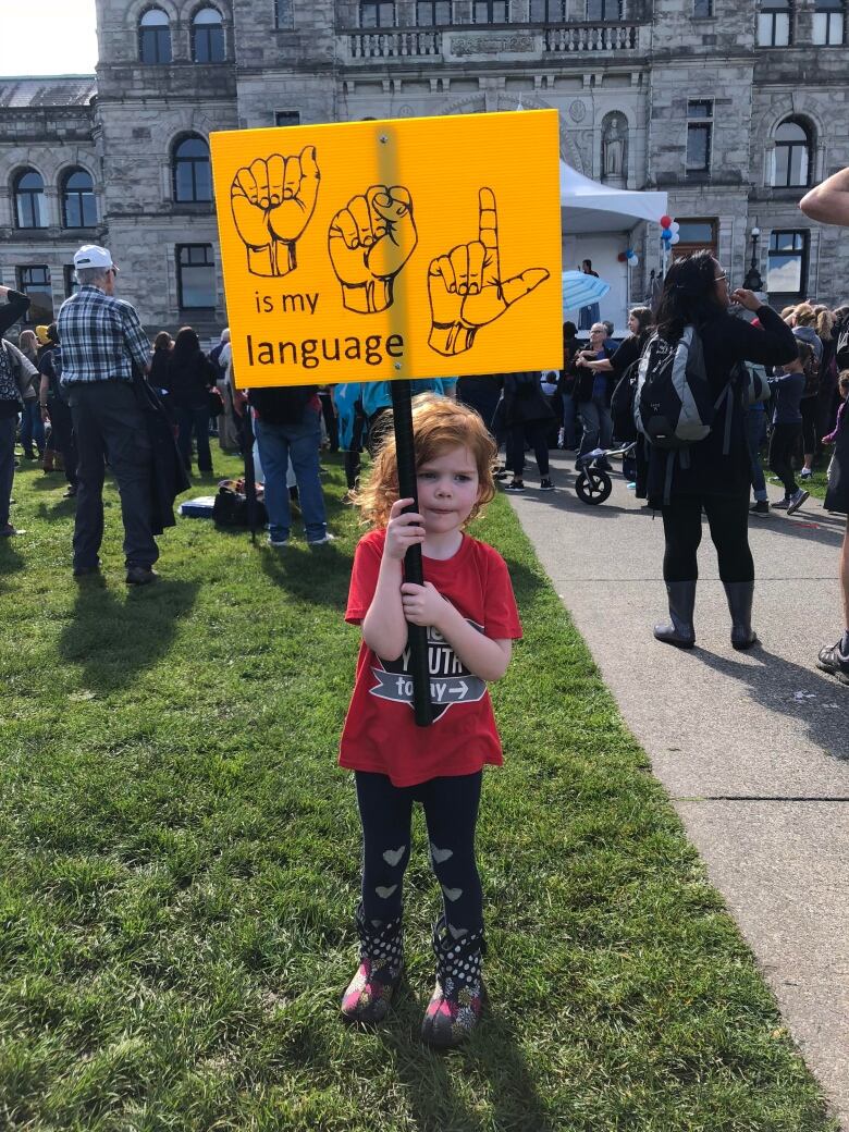 A girl in a red t-shirt and black leggings is pictured on a grass lawn, holding up a yellow sign that reads, 