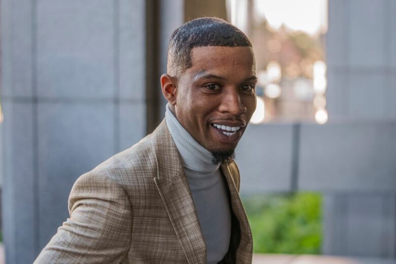 A man wearing a gray turtleneck and lightly coloured plaid jacket smiles.